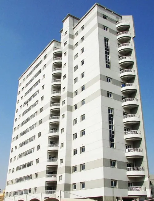 A tall, modern multi-story apartment building with balconies on one side, set against a clear blue sky.
