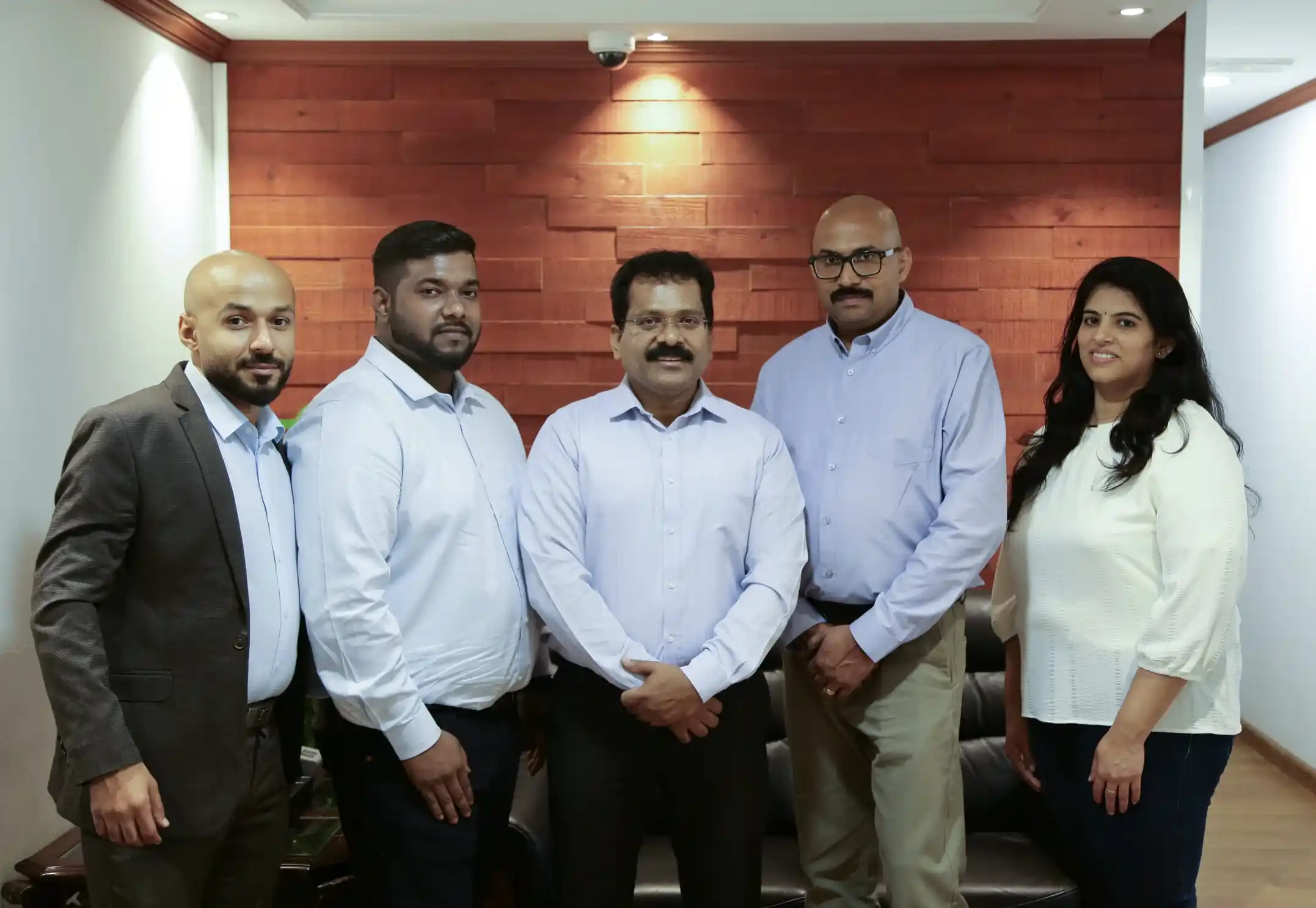 Five people are standing indoors with wood-paneled wall and recessed lighting behind them. They are dressed in business casual attire, and some are smiling faintly. Three men are on the left and two men and one woman are on the right