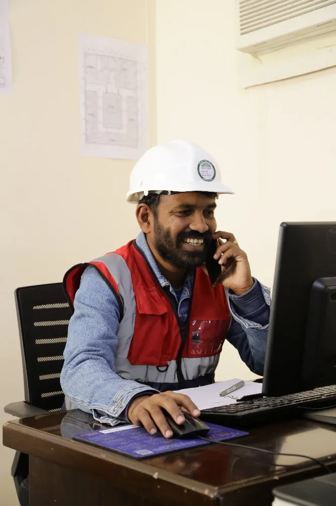 A man in a hard hat and vest sitting at a desk with a laptop, focused on work.