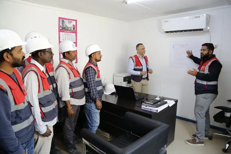 A group of six construction workers, five of whom are standing and one who is seated, are gathered in an office. They are wearing safety helmets and reflective vests. One person is pointing to a blueprint on the wall, possibly leading a discussion or briefing.