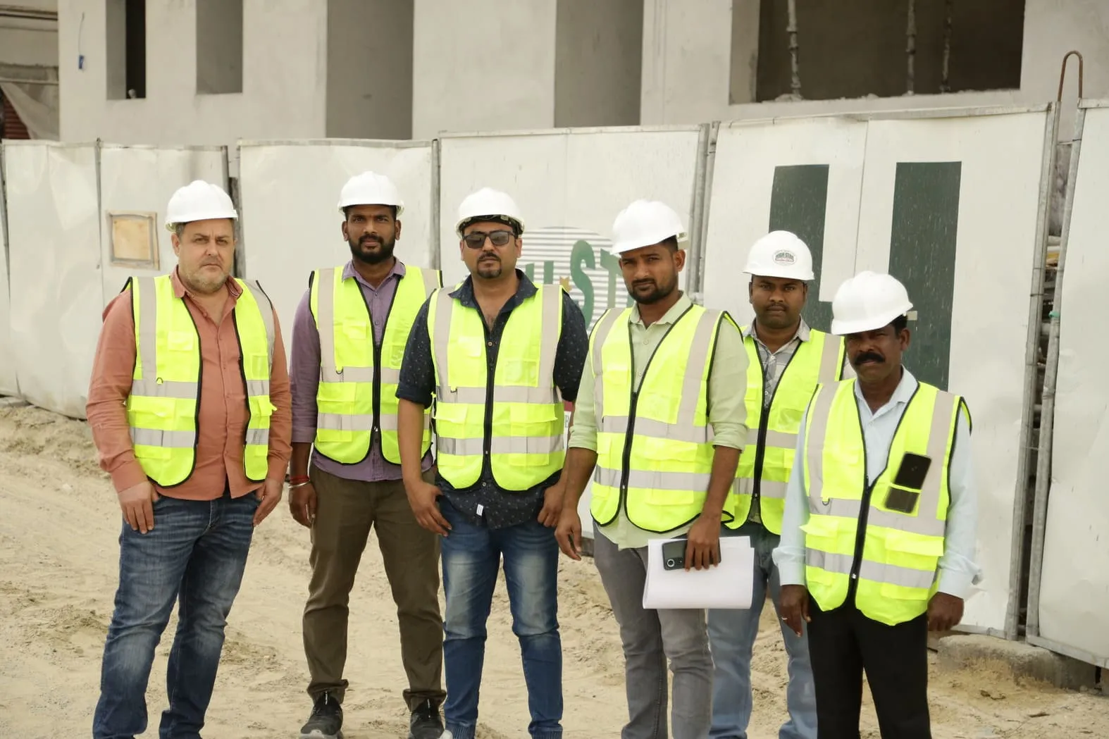 A group of six men wearing high-visibility vests and white hard hats stands in front of a construction site.