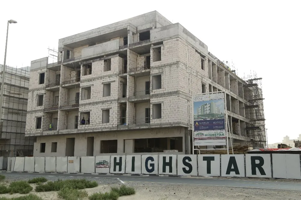 A multi-story building under construction with scaffolding on the right side. The facade is partially completed. There is a large sign with the word "HIGHSTAR" in front of the building. Construction boards and a billboard with a building image are visible.