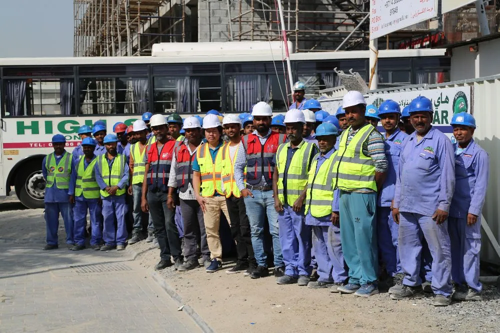 A group of construction workers in safety vests and helmets stands in front of a construction site, with scaffolding and a bus in the background.