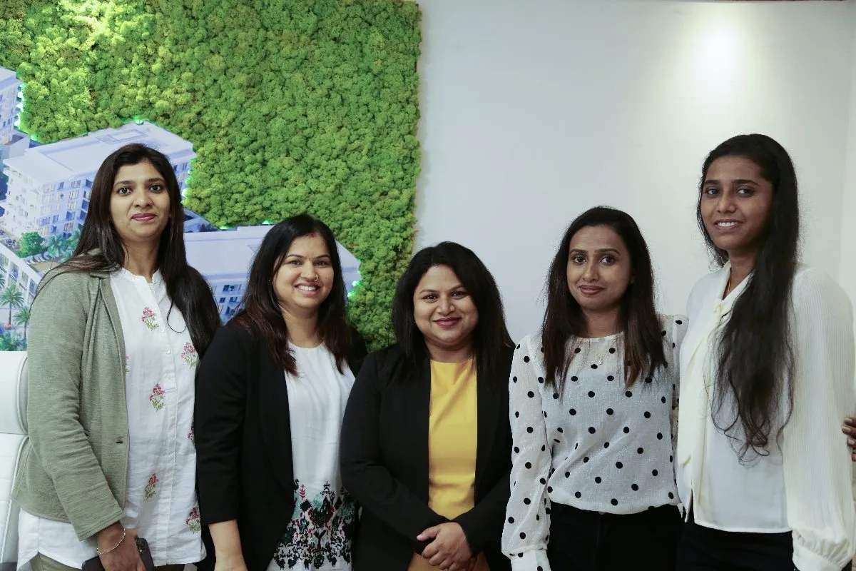 A group of five women stands together in front of a wall with green foliage and a cityscape map. They are all smiling and dressed in business casual attire.